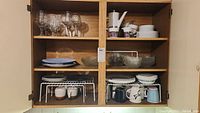 Cabinet shelves containing various plates, mugs, glasses, bowls, and a white ceramic tea set with floral decoration.