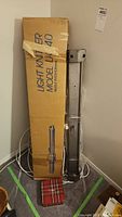 Original cardboard box labeled 'Light Knitter Model LK 40 Needle Pitch 6.5mm' leaning against wall next to metal stand for knitting machine.