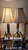 Two desk lamps side by side on a black metal stand with beige fabric shades and wooden bases with brass bottoms.