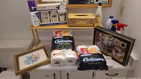 Overview of lot including skin care products, toilet paper, tissue box cover, framed wall art, and household items