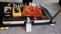 Photo showing Ouija board box, tarot card decks, and tarot books stacked on glass table.