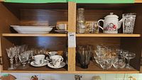 Wide view of wooden cabinet shelves holding glassware and tea sets including tea cups, saucers, dessert bowls, glasses, and teapot.