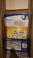 Stacked boxed twin window fans from Likewise, Sunbeam, and Comfort Zone, showing brand and product packaging.