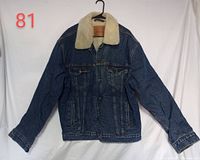 Front view of the blue denim Levi Strauss jean jacket with cream wool collar, hanging on a black hanger against a white background, showing front button closure and chest pockets.