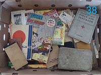 Photo showing a box containing various vintage paper ephemera including the 1940s First Aid textbook, colorful advertising brochures, and other booklets including the Cubby in Wonderland revised edition.