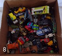 Top-down photo showing a cardboard box filled with a variety of small toy cars including cars, trucks, and utility vehicles in multiple colors and sizes.