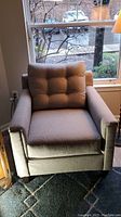 Front view of beige fabric armchair with attached tufted cushion positioned near window with greenery visible outside.
