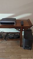 Photo showing the Bose AV3-2-1II media center unit on a wooden table, with remote control placed on top, and cables along with the subwoofer on the floor next to the table.