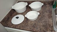 Set of three white CorningWare dishes and one matching lid with black knob sitting on countertop.