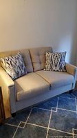 Full view of the beige Wayfair love seat showing tufted back cushions and two patterned pillows in a corner of the room on carpet