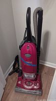 Full view of the vacuum cleaner standing upright against a wall, showing main body, handle, and attached extension hose.