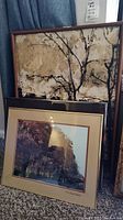 Three framed wall art pieces stacked upright on carpet: One large Michel de Gallard print with abstract cityscape and two framed photographs, one showing trees in autumn colors.