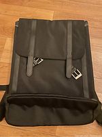 Front view of black backpack showing flap with two vertical straps with silver buckles, black fabric body, and padded bottom.