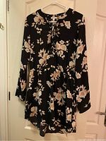Full front view of the black long sleeve dress with beige floral print hanging on a hanger against a cream door.