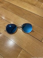 Top down photo of round sunglasses with blue mirror lenses and yellow gold metal frame on a wood floor.