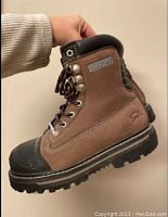 Side view of brown Dakota steel toed work boot held in hand showing genuine suede upper, black rubber toe cap, and branding on side