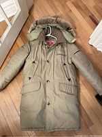 Full front view of the olive green Deacon Brothers down parka with fur hood laid flat on a wood floor.