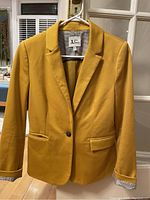 Front-facing image of a men's mustard yellow blazer on a hanger with a single button and two front flap pockets.
