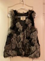 Front view of the faux fur vest showing patchwork pattern in blacks, greys, and browns, sleeveless with V-neck and hanging on a white hanger.