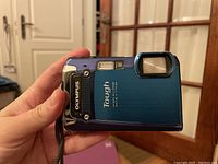 Blue Olympus Tough digital camera held in hand showing front and side, displaying model and brand logo.