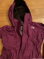 Front view of the purple The North Face women's waterproof jacket laid flat on a wooden floor showing hood and zippered pockets.