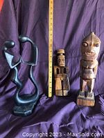 Photo showing the three figurine carvings together against purple backdrop with ruler height reference. Includes one modern abstract black stone carving and two wooden tribal carvings.