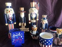 Four wooden nutcrackers with patriotic American flag-themed paint, two plaster bears in red, white, and blue clothing, and star-spangled tin containers for 4th of July decoration.