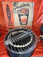 Photo of the metal round grill placed in front of the original box showing the cooking grate and metal handles.
