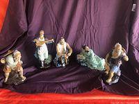 Photo showing all five ceramic Asian figurines arranged on a purple draped backdrop, showing full figures and some accessories missing.
