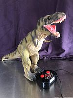 Front-side view of large plastic dinosaur toy standing upright with mouth open, beside black wired remote control with red buttons and Jurassic Park logo.