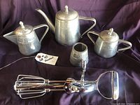 Full set showing pewter tea pot, creamer, sugar container, smaller pewter container, and metal egg beater on dark background.
