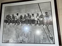 Framed black and white photograph print showing eleven factory workers sitting on a steel beam high above New York City during the construction of Rockefeller Center in 1932. The urban skyline is visible below them.