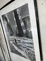 Framed black and white photographic print featuring surreal image of man walking through waves inside building with escalators.