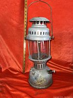 Full view of the vintage miners gas lamp showing metal body, glass enclosure, vent holes, and metal handle against a red fabric background with a measuring tape indicating about 16 inches in height.