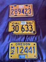 Front view showing New York (1975), California (1956), and Oregon (1976) vintage motorcycle license plates arranged on fabric background.