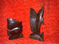 Photo showing both wooden carved sculptures: marlin with raised sail and fish with large dorsal fin, dark polished wood, placed on red background.