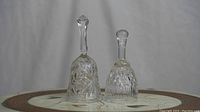 Two clear crystal bells with different cut patterns and handles placed on a round table with a white and brown inlay.
