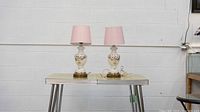 Side view of two white ceramic lamps with floral designs and pink shades placed on a small table against a white wall.