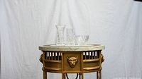 Three crystal glass items on round table with marble top and gold base, including a tall vase, medium tumbler, and short bowl, all clear with intricate cut patterns.