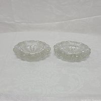Pair of clear crystal ash trays viewed from above, showing scalloped edges and circular indentation in center.