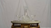 Glass decanter and three etched glasses on a round decorative wooden table with a white backdrop.