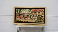 Front view of framed tapestry mounted on a stand showing street scene with horse-drawn carriage and house.