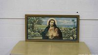 Framed religious print depicting Jesus Christ with glowing heart in outdoor setting inside wooden frame, shown on padded surface.