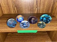 Six art glass paperweights arranged on a wooden shelf showing different internal designs and colors.