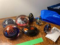 Seven assorted art glass paperweights arranged on wooden surface with a note and a green strip of tape
