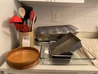 Photo showing Woodbury handcrafted wooden bowl, ceramic utensil holder with kitchen utensils including spatulas and spoons, and various baking pans and trays stacked.