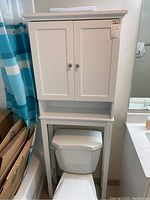 Full view of white bathroom storage cabinet mounted over toilet with box storage next to bathtub and blue shower curtain visible.
