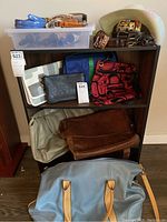 Photo of a black small leather clutch handbag, a red and black patterned handbag, a blue and green fabric bag, a beige handbag, brown suede shoulder bag, and a light blue handbag with tan handles placed on a wooden shelf. Above the shelf is a white cowboy hat and plastic bin with blue lint rollers and belts.