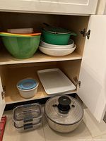 Cupboard shelf with three nested green bowls, white ceramic rectangular platter, green colander, white ceramic bowl with blue rim and lid, two plastic storage containers, and OXO salad spinner.
