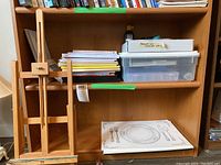 Wooden easel, books, papers, and white plastic container with calligraphy supplies inside a wooden shelf